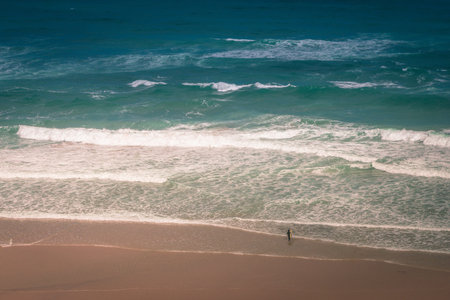 View of the Guincho beach near Atlantic coast. Surfer on the ocean coast in a wet suit with surfboard. Landscape of sunny day, blue sky and a mountain in distance. Cascais. Portugal.の写真素材