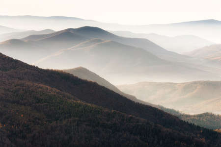 Beautiful landscape of autumn mountains layers range at hazy morning. A view of the misty slopes of the mountains in the distance and forest hills in rays of sunlight.の写真素材