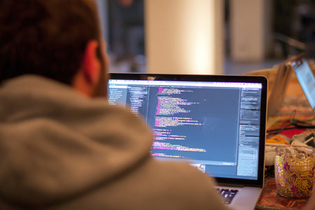 Over-the-shoulder view of a programmer's desk with an open laptop and code being typed on the screen. A programmer is writing software code by hand.の素材