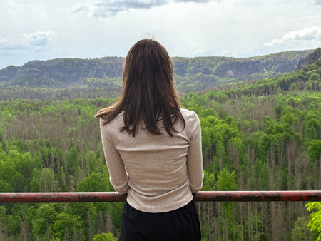 A young woman admires the view of forested cliffs from the observation deck at the summit. Hiking. Rear view of a woman in the mountains on a cloudy day in spring.の写真素材