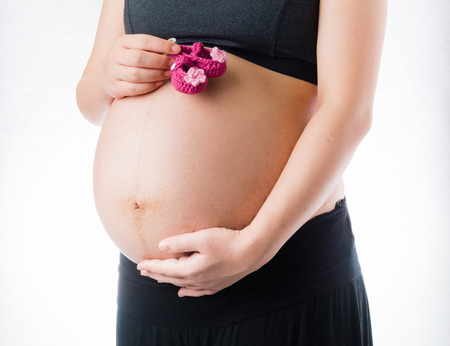 Happy pregnant woman take pink wool baby shoes in hand and touch belly, isolated on white backgroundの写真素材