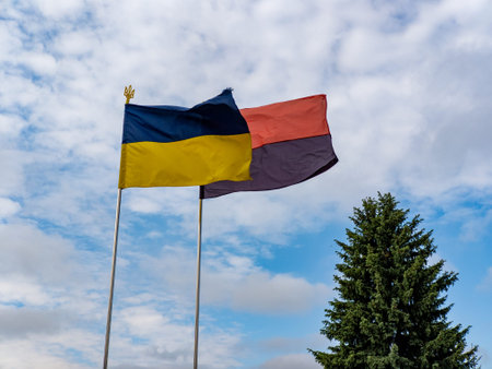 Two flags flying in the wind against a cloudy blue sky.
