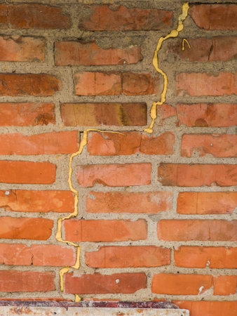 Close-up of a weathered red brick wall featuring a prominent vertical crack filled with yellow...