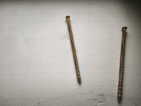 Close-up of two brass-colored screws lying on a light gray concrete wall with visible texture and minor cracks. The screws have threaded shafts and rounded heads, showcasing industrial precision against a rough, neutral backdrop. Ideal for construction, hardware, or industrial-themed projects.の写真素材