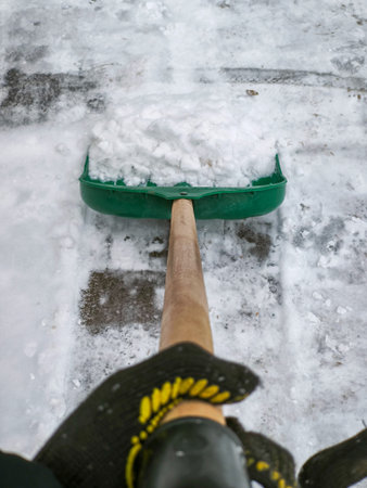Close-up view of a person shoveling snow with a green plastic shovel. The snow is packed and freshly cleared, revealing patches of icy ground. The wooden handle is held firmly, with a gloved hand visible. Focus on the effort of winter cleanup.の写真素材