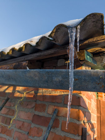 Close-up of long icicles hanging from a snow-laden roof edge. Bright sunlight highlights the clear...
