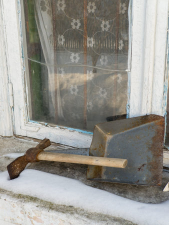 A rusty, vintage shovel with a wooden handle rests beside a corroded metal container on a snowy porch. Behind them, a weathered window with frayed floral curtains frames the scene, evoking nostalgia and abandonment. The cold winter atmosphere highlights the contrast between decay and remnants of domestic life.の写真素材