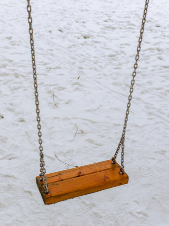 A solitary wooden swing, suspended by metal chains, hangs empty in a snow-covered playground. The ground is blanketed in fresh snow, with faint footprints and scattered twigs. Contrasting the warm wood against the cold, white landscape, this serene winter scene evokes quiet stillness of a frozen day at the park.の写真素材