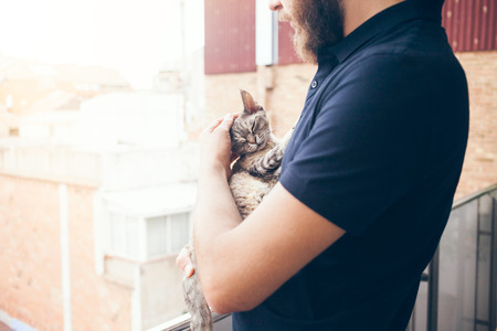Young man is standing on a balcony with his Devon Rex cat. Home pets. Embracing, love, lifestyle. Cat peopleの写真素材