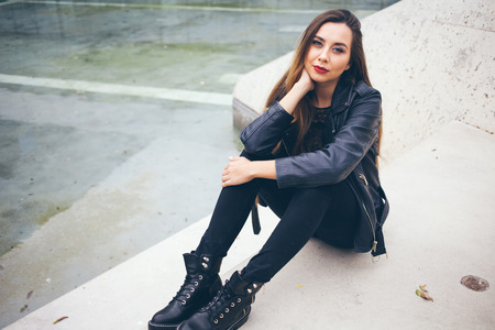 Beautuful young teenage girl is sitting on the concrete steps. Student in alternative style clothes - black leather jacket and big boots. Copy-space area for your advertisement text or designの写真素材