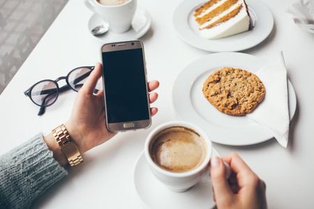 Close-up of woman's hand holding cell phone while drinking coffee and eating oat cookie. Reading news, surfing the web / internet,chatting in social network  in smartphone. Blurred background.の写真素材