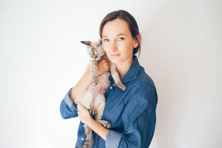 Lifestyle photo of a woman in casual clothes holding and hugging cute sleepy Devon Rex kitty. Cat is...