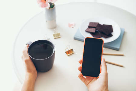 Close-up of woman's hands holding cell phone while drinking tea and eating chocolate. Reading news, surfing the web / internet, chatting in social network in smartphone. Blurred background.の写真素材