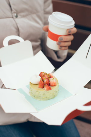 Close-up of woman hang holding cup of take away coffee and going to eat pistachio pastry flavored with fresh strawberries gluten free and vegan in box to go. Natural light, selective focus photoの写真素材