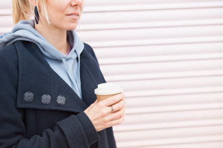Close up photo of a young blond woman in casual clothes holding and drinking cup of coffee. Pale...