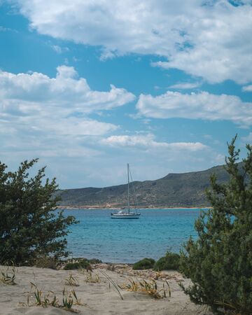 Sailboat seen from between green bushes in Elafonisos, Greece on a beautiful day with blue sky and clouds.の写真素材