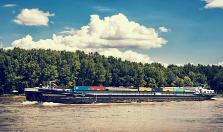 Cargo ship with containers on the Danube river, Slovak republic. Goods transport. Blue retro photo filter. Freight transportation. Against the flow. Clouds in the sky.の写真素材
