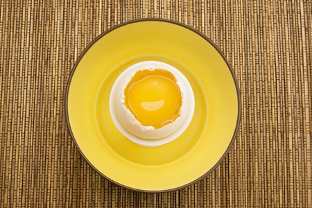 Raw egg cracked open with yellow yolk inside a white eggshell in a yellow bowl on top of a bamboo...