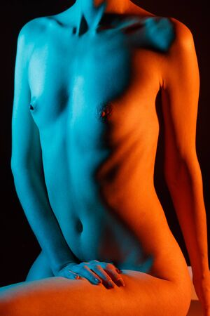 A beautiful naked girl in a blue-orange light without a face sits on a cube, her right hand on her hip, sowing from behind.の写真素材