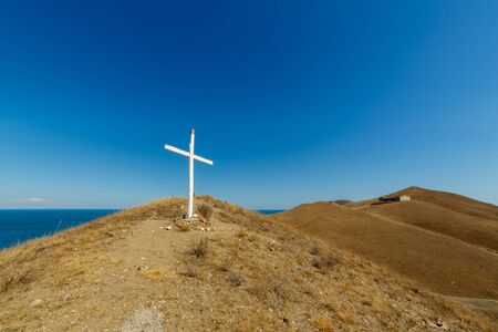 Christian wooden cross on mountain top, rocky summit, beautiful inspirational landscape with ocean, island, clouds and blue sky, looking at scenic blue sea and white clouds.の写真素材
