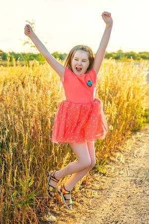 Cute screaming little girl in pink dress jump outdor in field in sunset. Activities with children summer outdoors. Having fun. Vertical.の写真素材