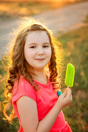 Cute smiling little curly girl in pink dress with green ice-cream in her arm outdor in sunset. Activities with children summer outdoors. Vertical.の写真素材
