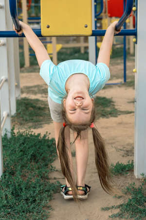 Cute little girl with two funny tails hanging upside down and having fun at playground. Activities with children summer outdoors. Vertical.の写真素材