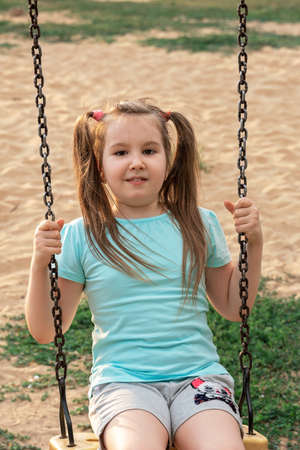 Adorable smiling little girl with two funny tails having fun on a swing at playground. Activities with children summer outdoors. Vertical.の写真素材