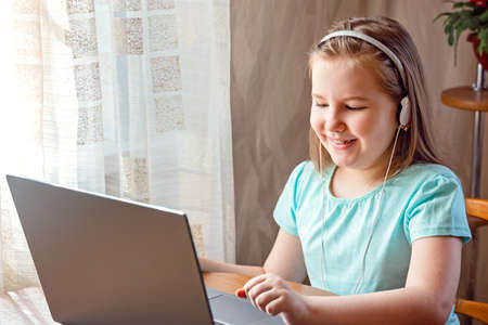 Distance learning online education. Happy smiling little girl in headphones is using a laptop and study online with video call teacher at home. New normal. Social distancing.の写真素材