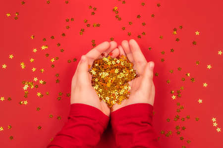 Kids hands in red blouse holding golden stars confetti on red background. Christmas, winter, new year and any holiday concept. Flat lay, top view.の写真素材