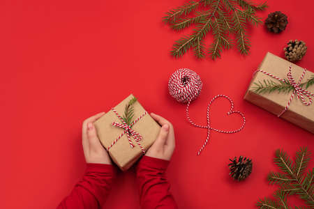 Girl hands holding present for Christmas. Packed gift in craft paper and scroll, spruce branches on red background. Workplace for preparing handmade gifts. Top view. Zero waste Christmas flat lay.の写真素材