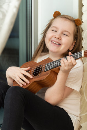 Portrait of happy smiling little girl in white t-shirt and black leggings sitting inside of home and playing ukulele. Hobbies and leisure activities concept. Vertical.の写真素材