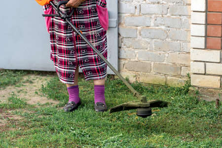 A senior women with a gas mower in her hands, mowing grass in front of the house. Process of mowing tall grass with a trimmer. Self-supply and housework concept.の写真素材