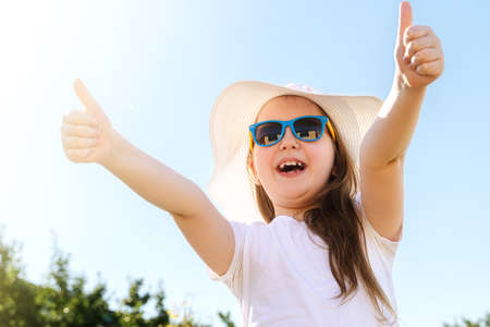 Happy smiling little girl in sunglasses, white t-shirt and summer hat against blue sky background. Summer vacation, travel and adventure concept.の写真素材