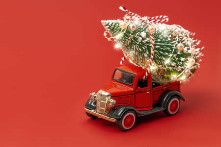 Red small retro toy truck with sparkling Christmas tree lights on truck body on red background. Delivery, christmas, New Year concept. Copy space, selective focus.の写真素材