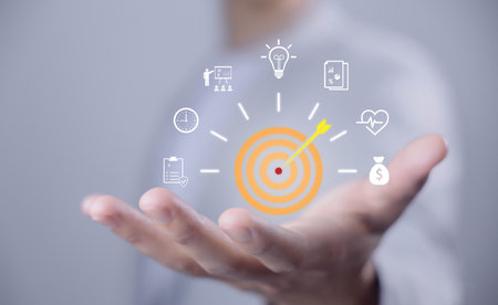 Goal setting and achievement concept. Hand holding a digital dartboard with an arrow hitting the target, surrounded by icons of time, money, health, ideas, planning, and success strategy.の写真素材