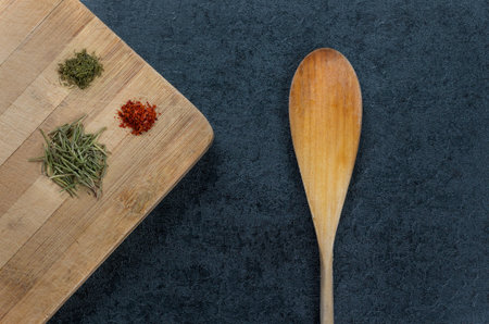 Spices such as dried rosemary plants, dills and red pepper on wooden chopping board with spoon on grayish groundの写真素材
