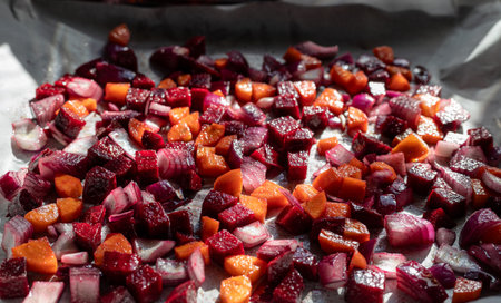 A baking tray with parchment paper holds diced beets, carrots, and red onions, ready for roasting. Sunlight highlights the vibrant colors and fresh ingredients.の写真素材