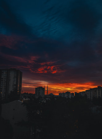 Dramatic morning light over an urban skyline with silhouetted buildings and mosque minarets. Deep blue clouds contrast with fiery orange sunrise tones, creating a moody, cinematicの写真素材