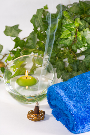 Lit candle with incense, some leaves and a blue towel, in a bathroom or a spa, with white backgroundの写真素材