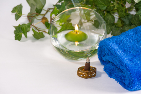 Lit candle with incense, some leaves and a blue towel, in a bathroom or a spa, with white backgroundの写真素材
