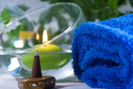 Lit candle with incense, some leaves and a blue towel, in a bathroom or a spa, with white backgroundの写真素材