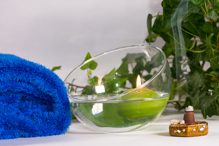 Lit candle with incense, some leaves and a blue towel, in a bathroom or a spa, with white backgroundの写真素材