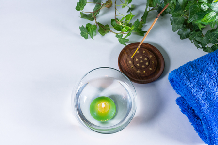 Lit candle with incense, some leaves and a blue towel, in a bathroom or a spa, with white backgroundの写真素材