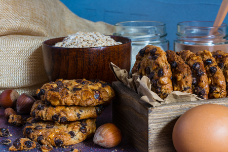 Box with oats cookies with hazelnuts and chocolate chips, a bowl with oats flakes, jars with chocolate and flour, some hazelnuts and one eggの写真素材