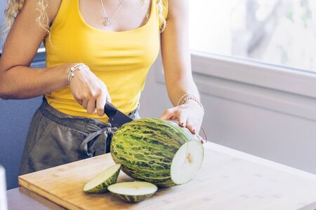 Young blonde girl cutting a melon with a knife, in the kitchen by a windowの写真素材