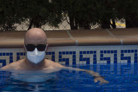 Adult man with sunglasses and medical mask bathing in a pool. Concepts like summer, end of quarantine, lockdown restriction, or new normalの写真素材