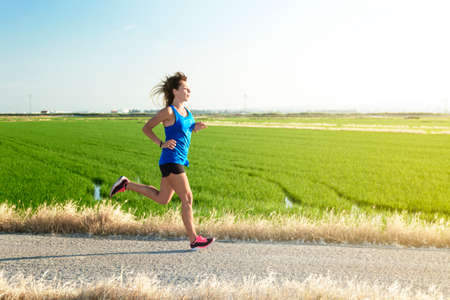 A young athlete girl in a blue shirt running down a path between rice fields, with her reflection in the paddy waterの写真素材