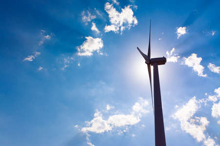 Backlight of a windmill tower with the sun behind the engine and cloudy blue sky with beams of sunlight. Clean energy, climate change and ecology conceptsの写真素材