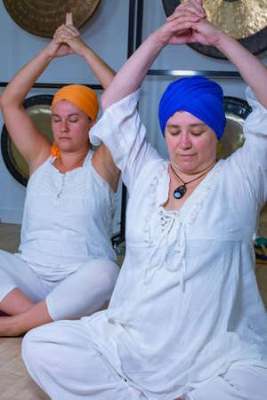Two women dressed in white with turban practicing kundalini yoga sitting on the floor with their hands on their headsの写真素材